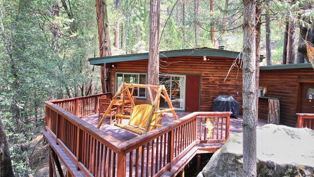 Una cabaña con terraza de madera, un conjunto de columpios y una gran roca al frente.
