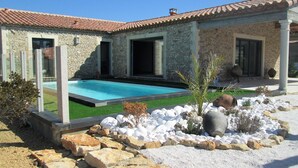 A stone house with a swimming pool, a patio with white pebbles, and a garden with rocks and plants.