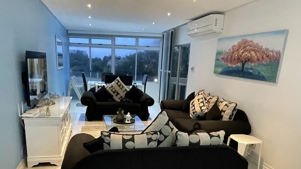 A modern living room with a black sofa, a white side table, and a glass coffee table. There is a painting on the wall and a large window providing a view of the outdoors. - A fantastic modern and spacious apartment right on Umhlanga main Beach . (Umhlanga)
