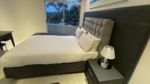 A bedroom with a large bed, a bedside table with a lamp, a window with a view of palm trees, and a painting on the wall. - A fantastic modern and spacious apartment right on Umhlanga main Beach . (Umhlanga)