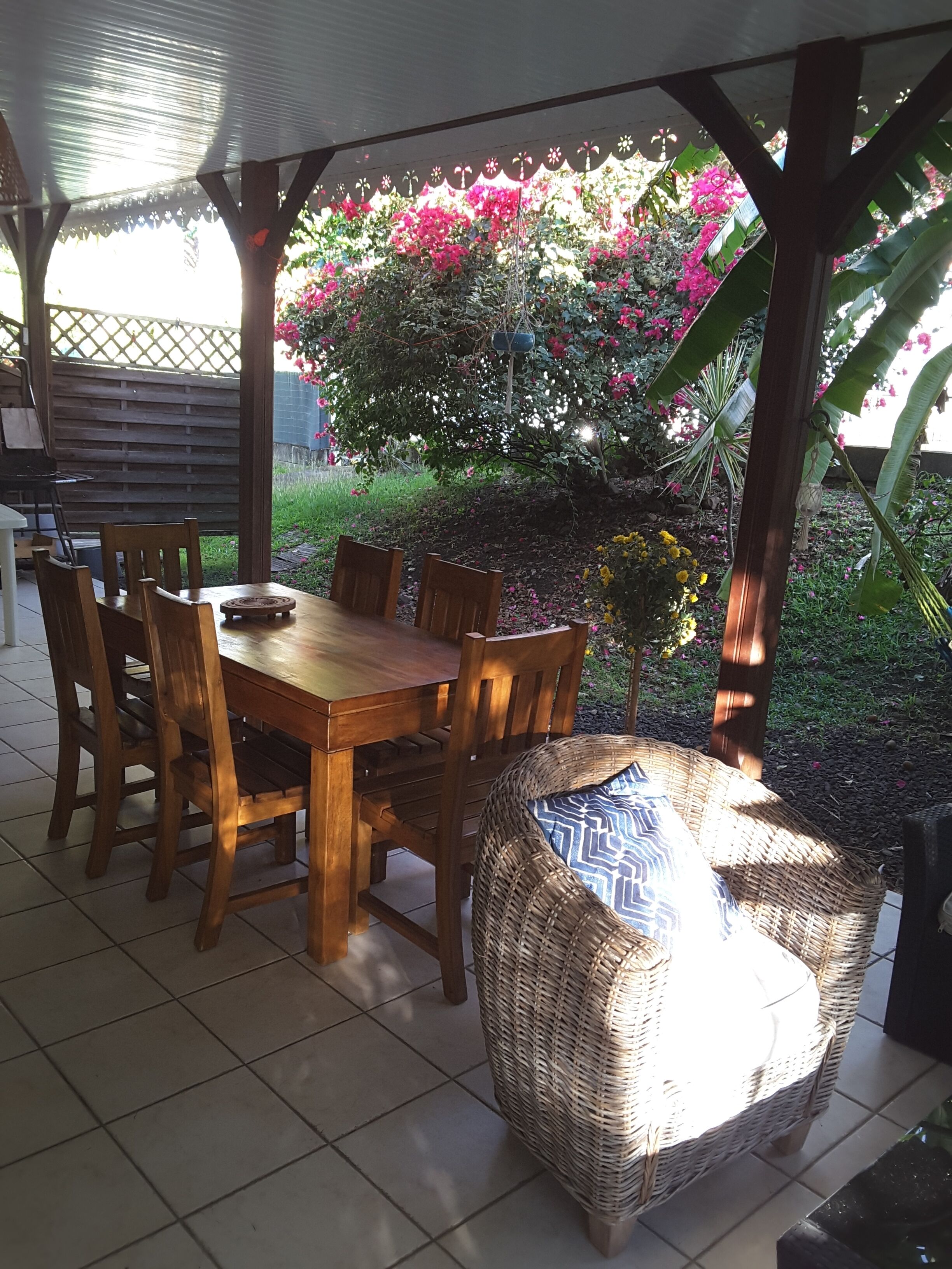 Une table de salle à manger en bois, des chaises, un fauteuil en osier et un patio couvert avec vue sur un jardin où poussent des fleurs roses.