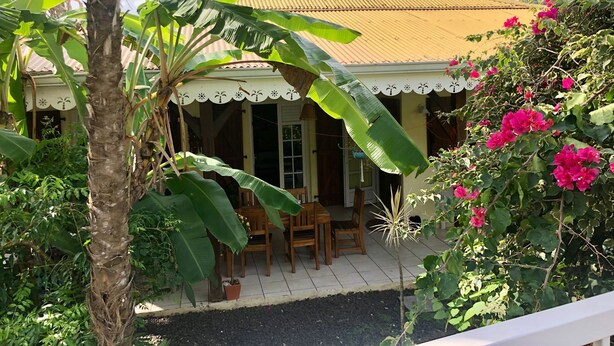 A house with a tiled patio, a covered outdoor seating area, and lush greenery including banana plants and bougainvillea flowers. - Villa with Caribbean Sea Small Pool (Les Trois-Îlets)