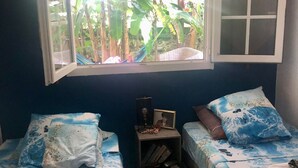 A bedroom with two beds, a window with a view of greenery, and a small table with a fan and picture frame. - Villa with Caribbean Sea Small Pool (Les Trois-Îlets)