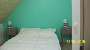 Une chambre avec un plafond en pente, des murs de couleur turquoise, un lit avec une literie blanche, deux tables de chevet avec des lampes et une prise électrique visible.