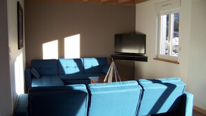 Un salon avec un canapé bleu, un téléviseur à écran plat fixé au mur et une fenêtre laissant entrer la lumière naturelle.
