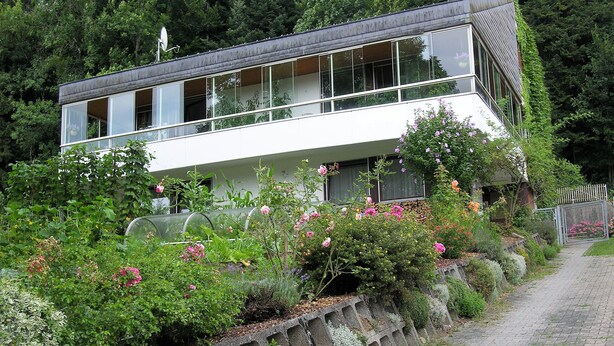 A modern two-story house with large windows and a balcony, surrounded by lush greenery and flowering plants. - Family-friendly apartment on the Hofsteig (Schwarzach)