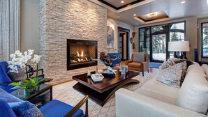 A modern living room with a stone fireplace, blue and white furniture, and large windows offering a view of the outdoors. - Ski-in/out Luxury Mansion, Game & Theater Rooms, Private Hot Tubs! Park City Colony Grand (Park City)