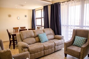 A living room with a beige sofa, two armchairs, a dining table with chairs, and a large window with curtains. - Zawiya property (Nairobi)