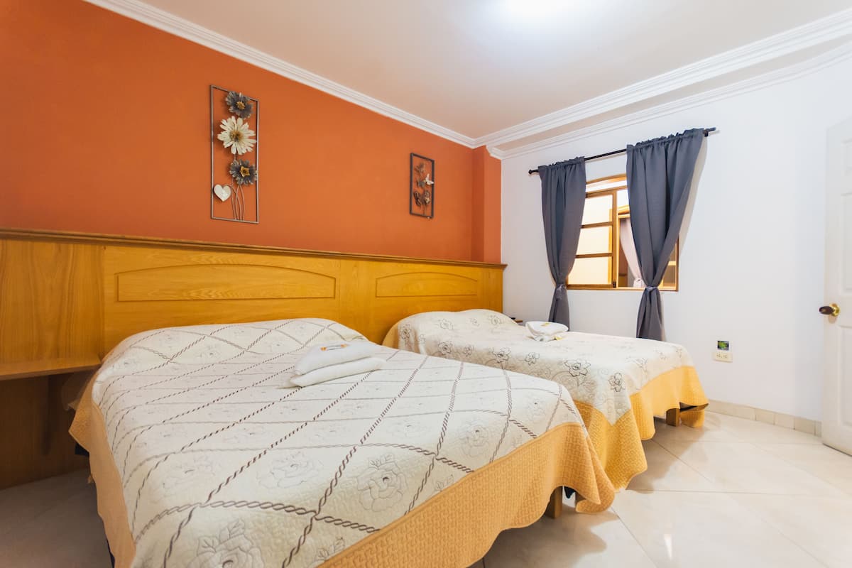 Quarto com duas camas, cabeceiras de madeira, uma janela com cortinas e uma porta.