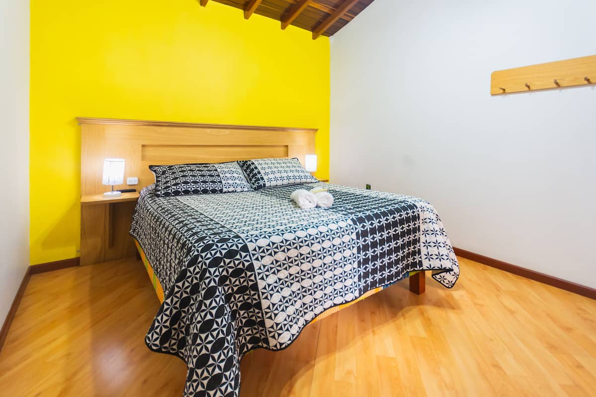 Uma cama com colcha estampada, luminária de cabeceira e cabeceira de madeira em quarto com paredes amarelas e piso de madeira.