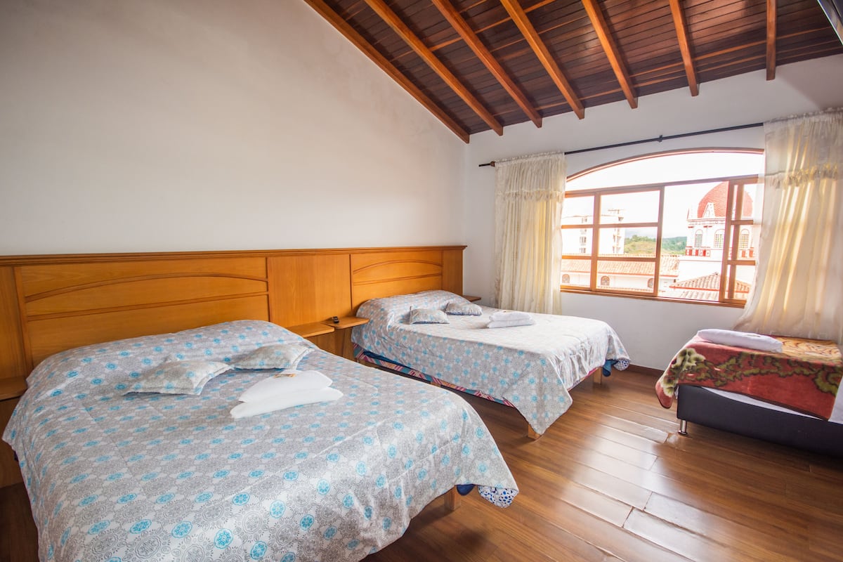 Quarto com duas camas, cabeceiras de madeira, piso de madeira e janela com vista para um edifício.