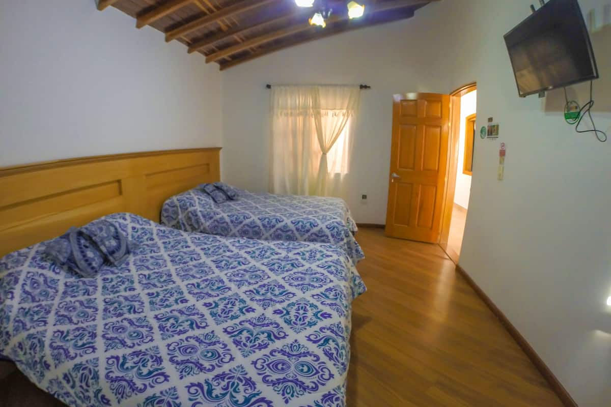 Quarto com duas camas, vigas de madeira no teto, televisão fixada na parede e porta que dá para outro cômodo.