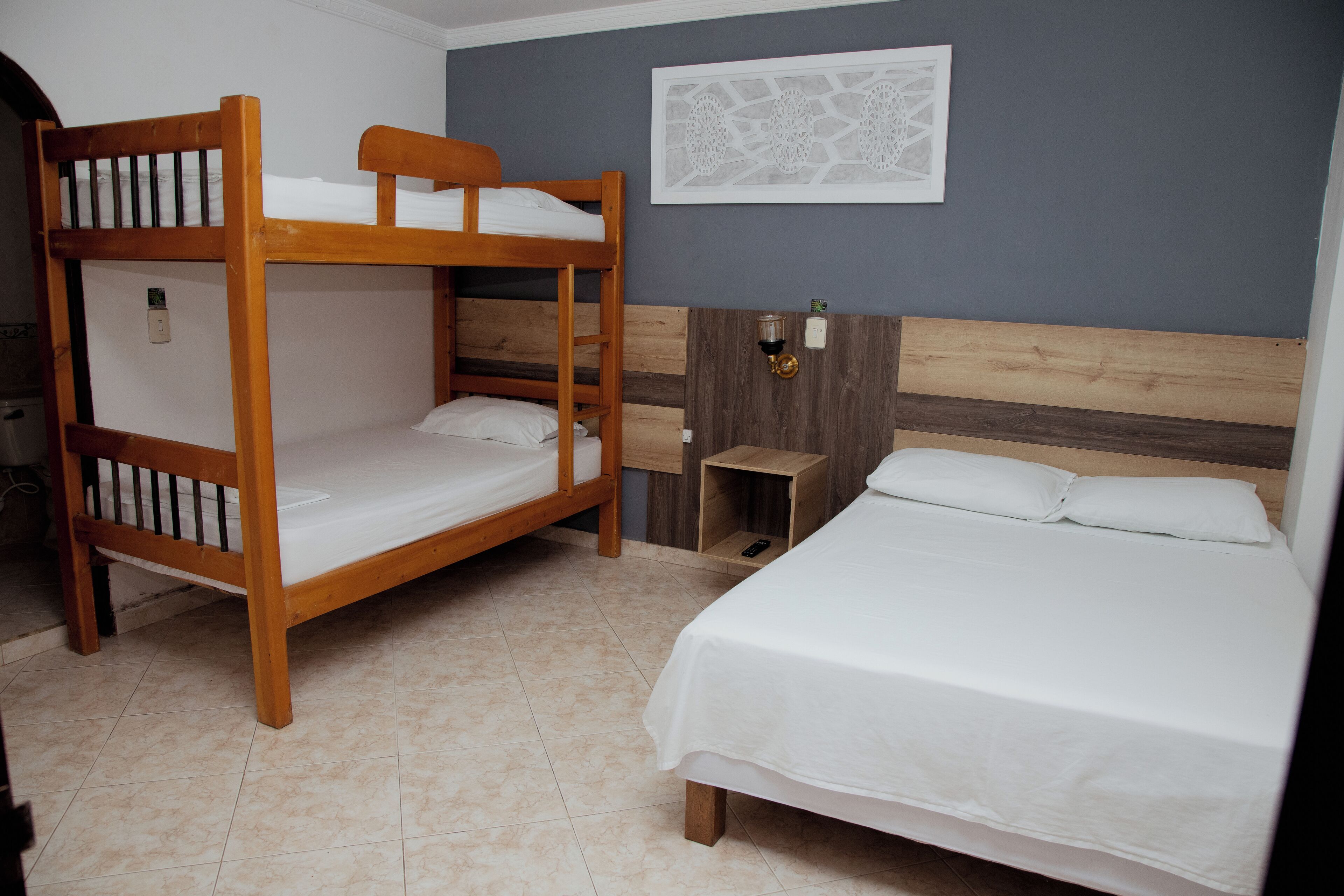 A room with two wooden bunk beds and a single bed, a small wooden nightstand, and a wall-mounted light fixture.