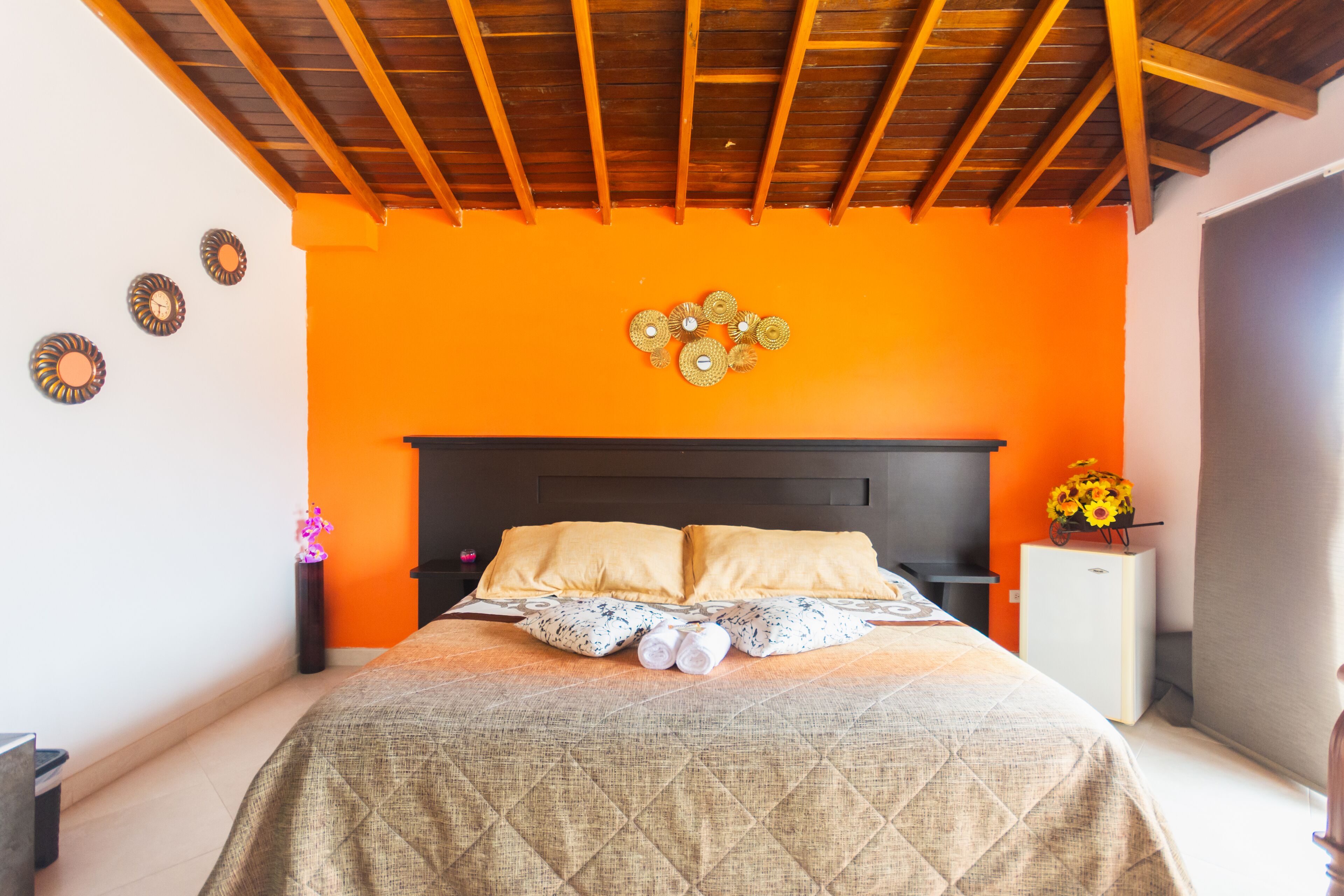 A bedroom with an orange wall, a wooden ceiling, a bed with two pillows, a nightstand, a small white cabinet, and a vase with flowers.