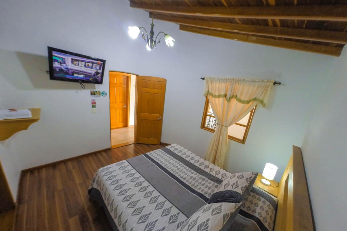 Quarto com cama, televisão na parede, teto de madeira e janela com cortinas.