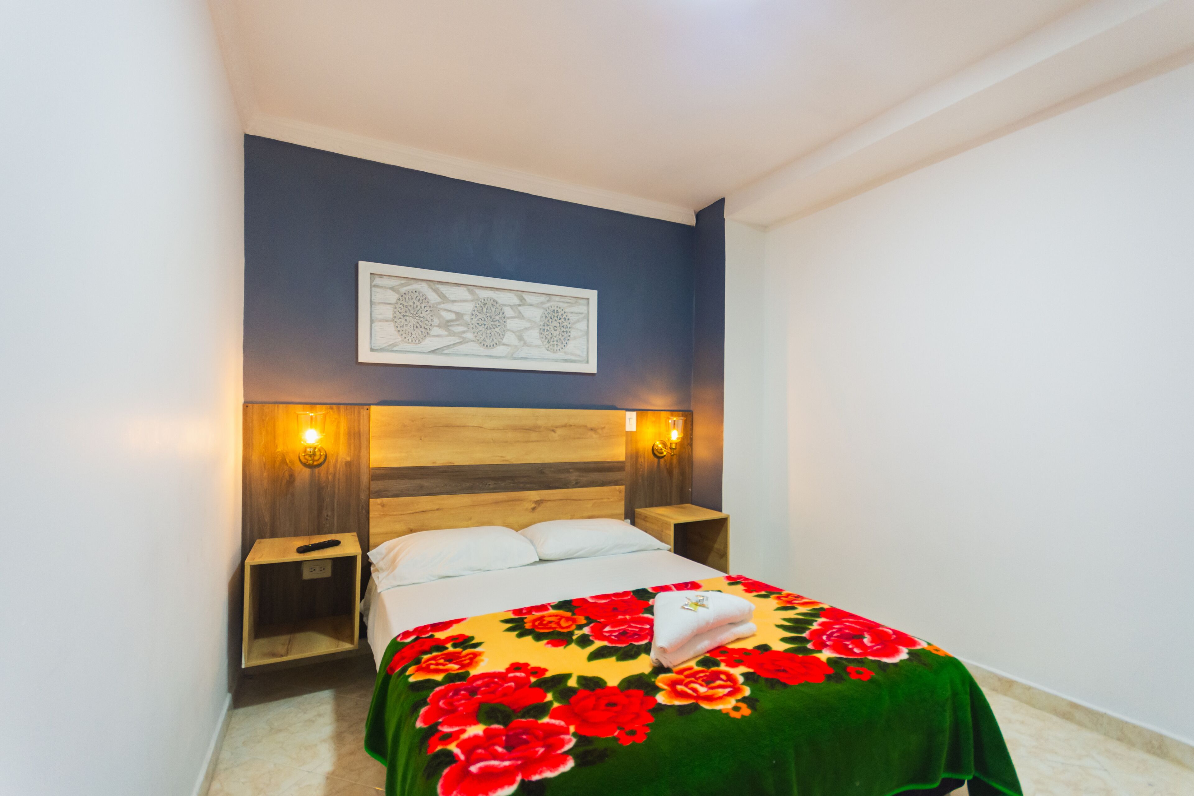 A hotel room with a bed, wooden headboard, two bedside tables, a wall-mounted light fixture, and a framed artwork above the bed.