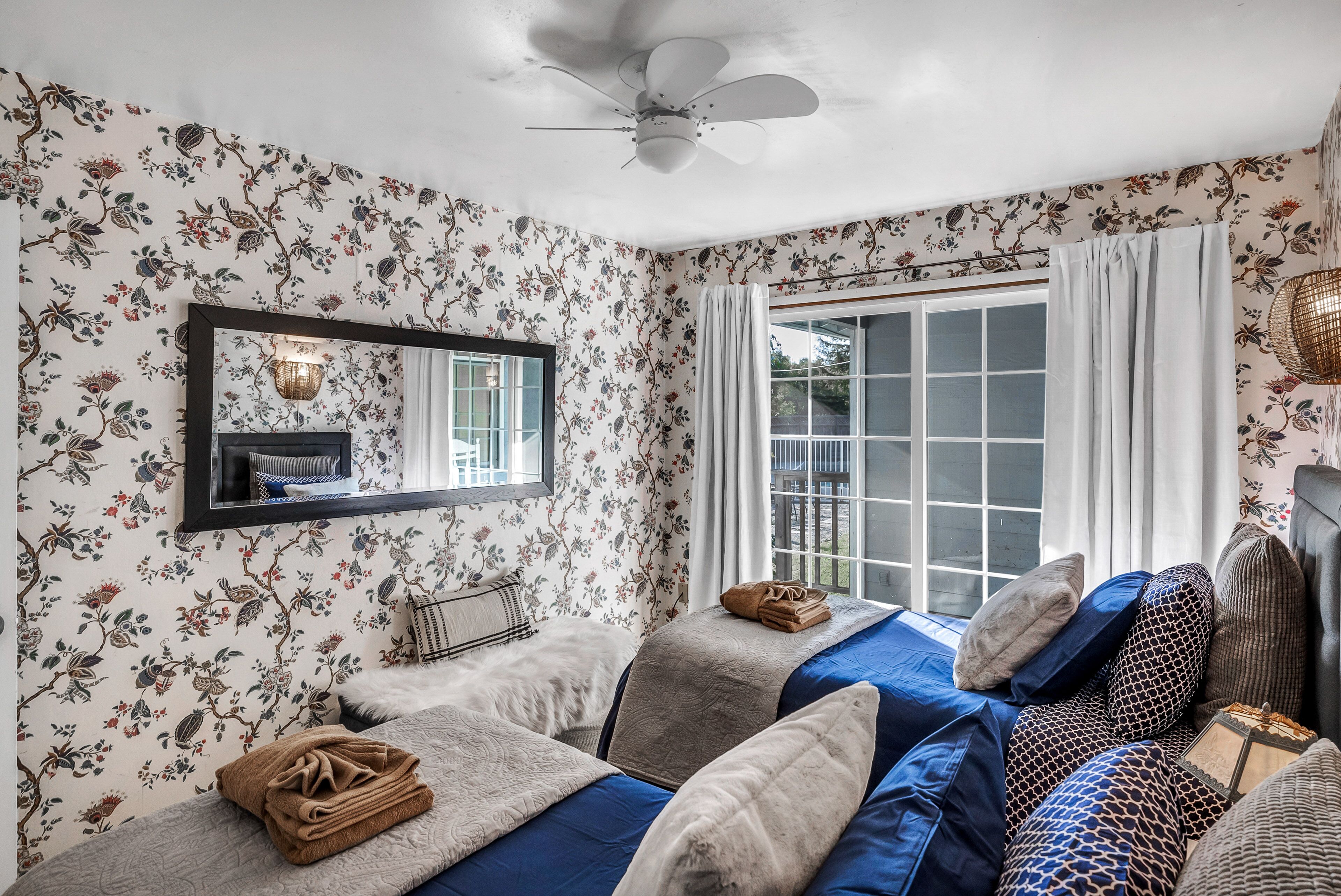 A bedroom with floral wallpaper, a ceiling fan, a large window with curtains, and a bed with multiple pillows and a blanket.