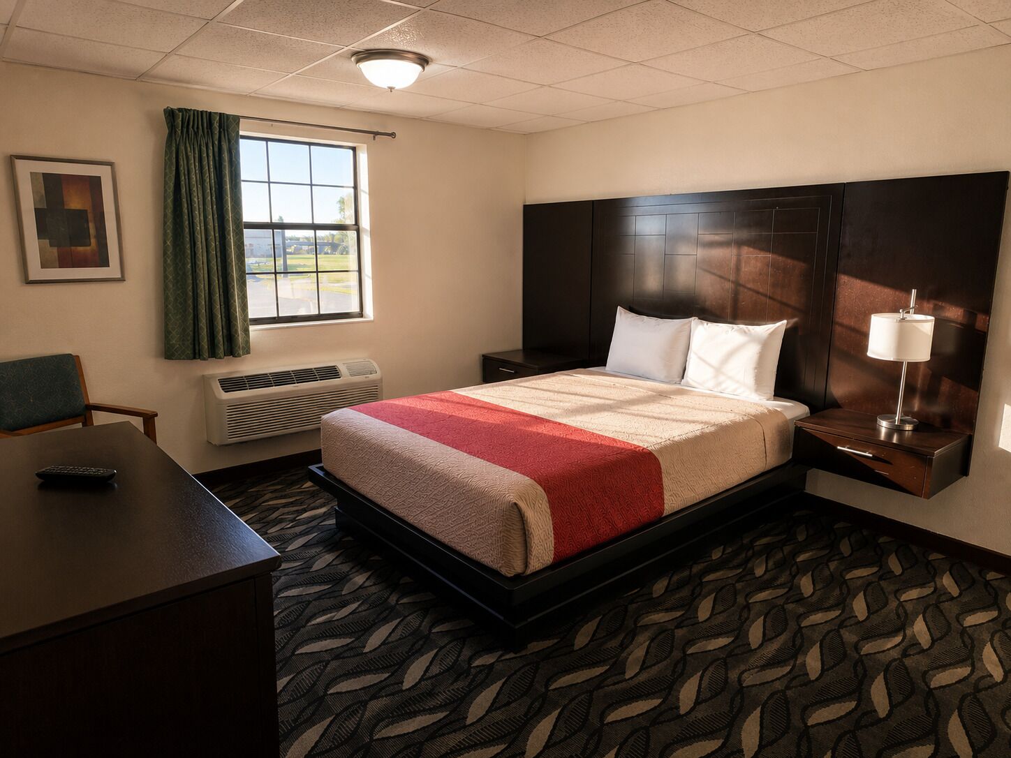 A hotel room with a bed, a desk, a chair, a window with curtains, an air conditioning unit, and a nightstand with a lamp.