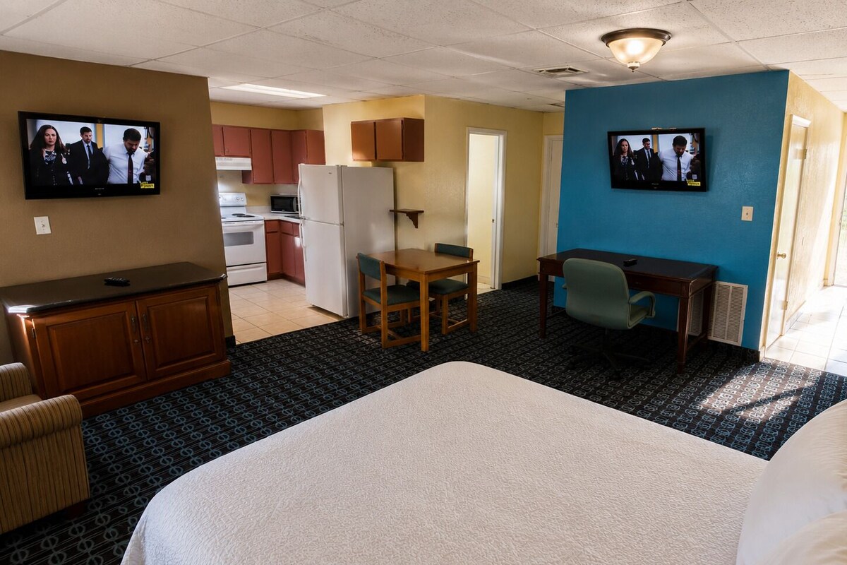 Quarto de hotel com cama, escrivaninha, cadeira, televisão e cozinha compacta.