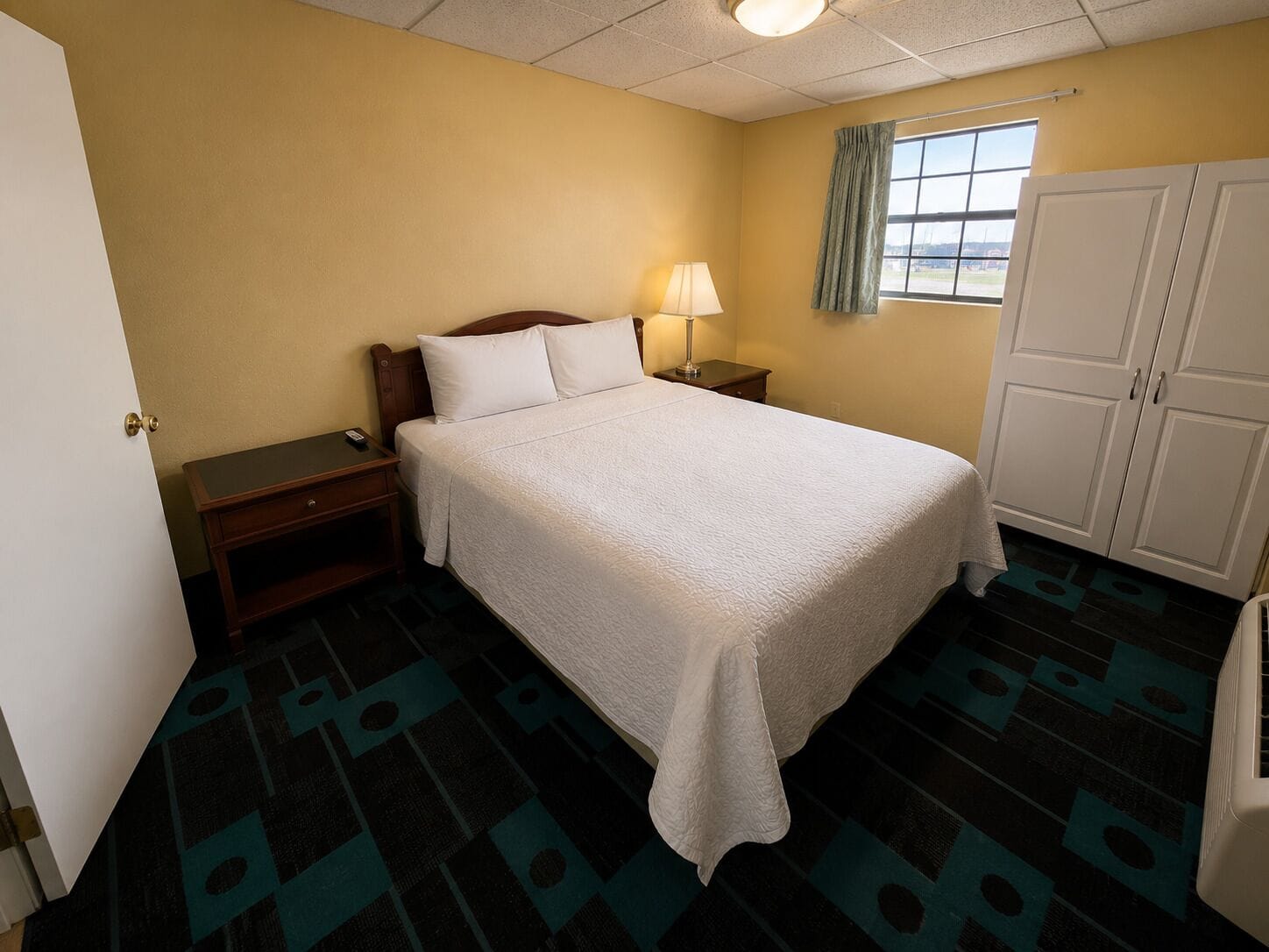 Una habitación de hotel con cama, mesita de noche, lámpara, ventana con cortinas, armario y alfombra estampada.