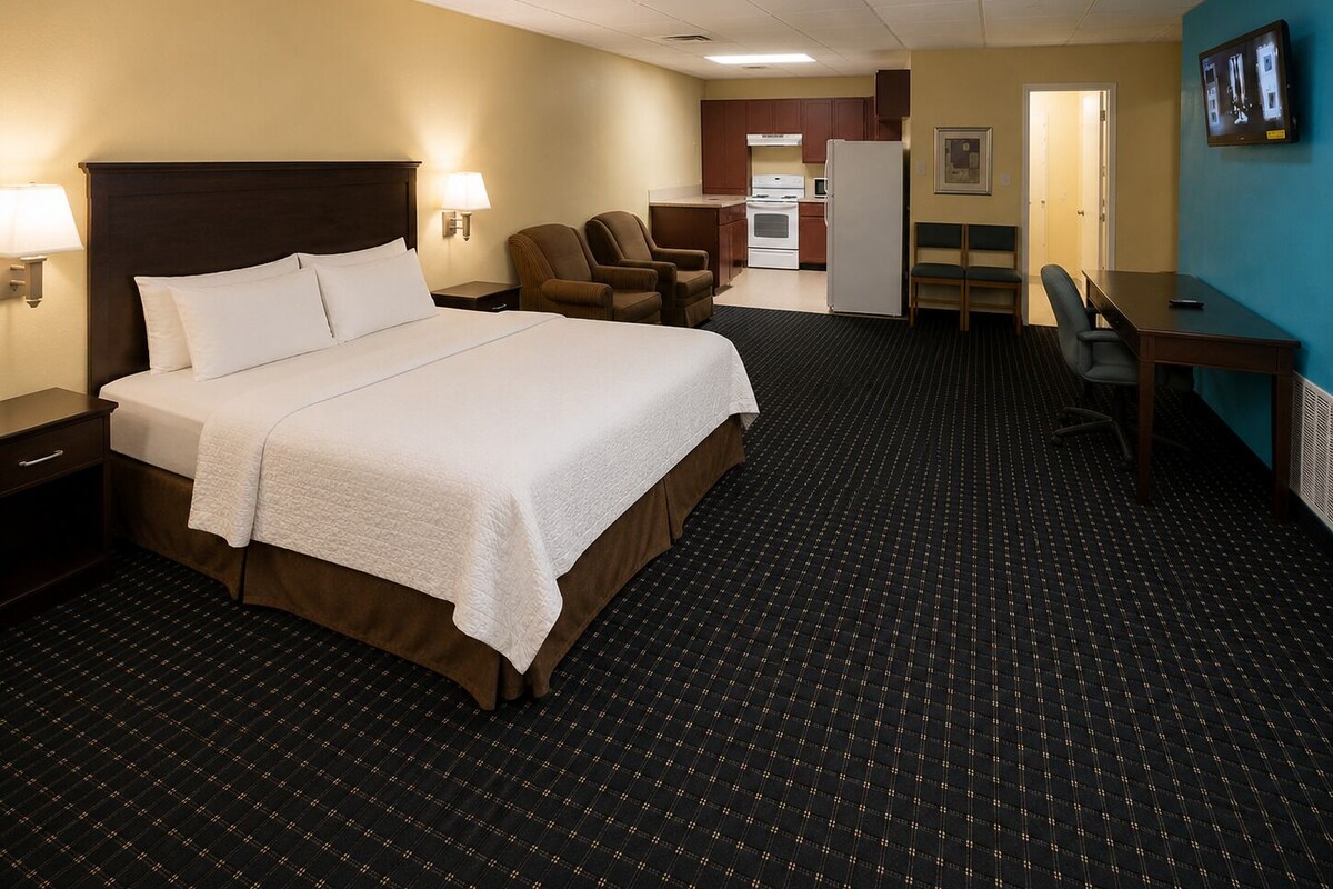 Quarto de hotel com cama, escrivaninha, cadeira, cozinha compacta e televisão.