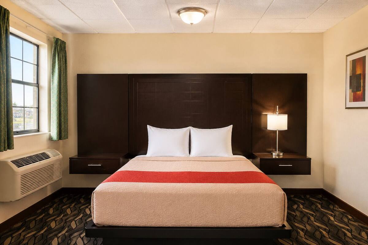 Une chambre d’hôtel avec un grand lit, deux tables de chevet, une lampe fixée au mur et une fenêtre avec des rideaux.