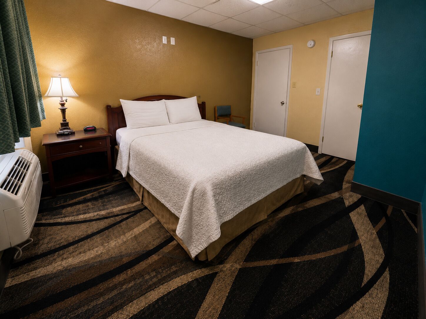 A hotel room with a bed, a nightstand, a lamp, an air conditioner, and a door.