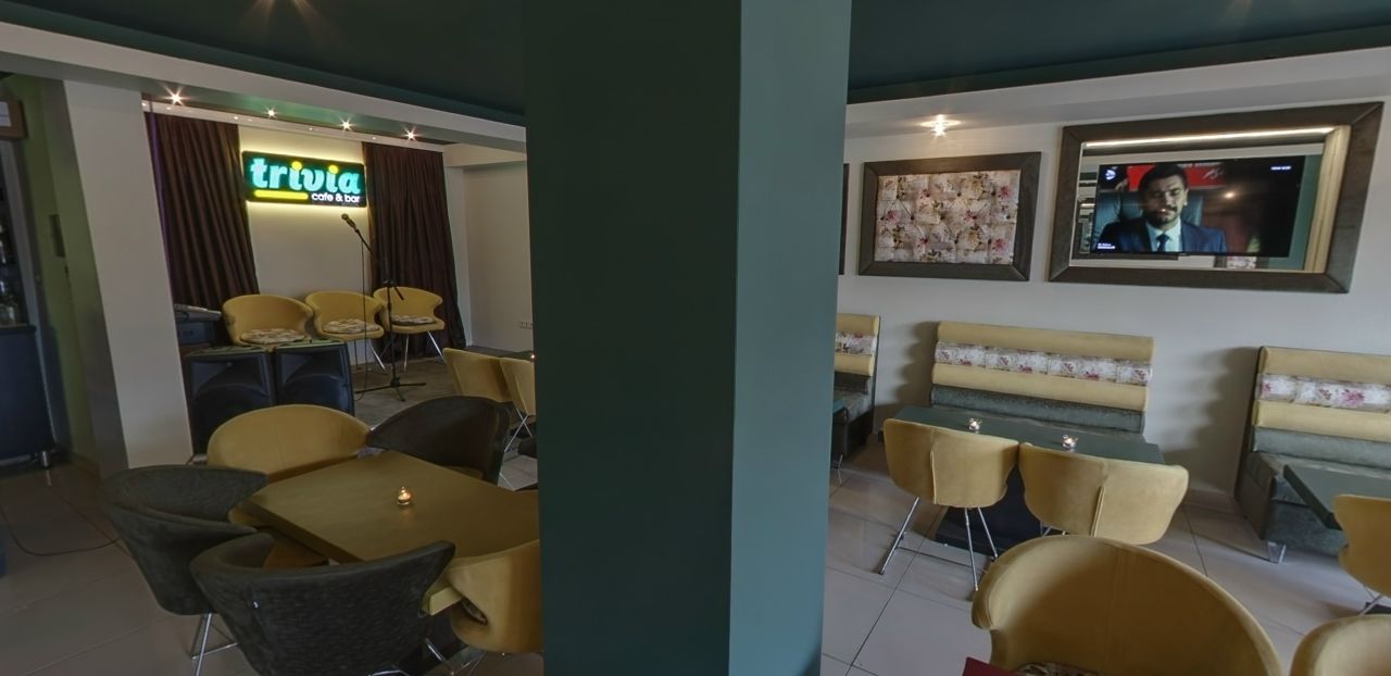 A restaurant with yellow chairs and tables, a 'Trivia cafe & bar' sign, and a TV screen on the wall.