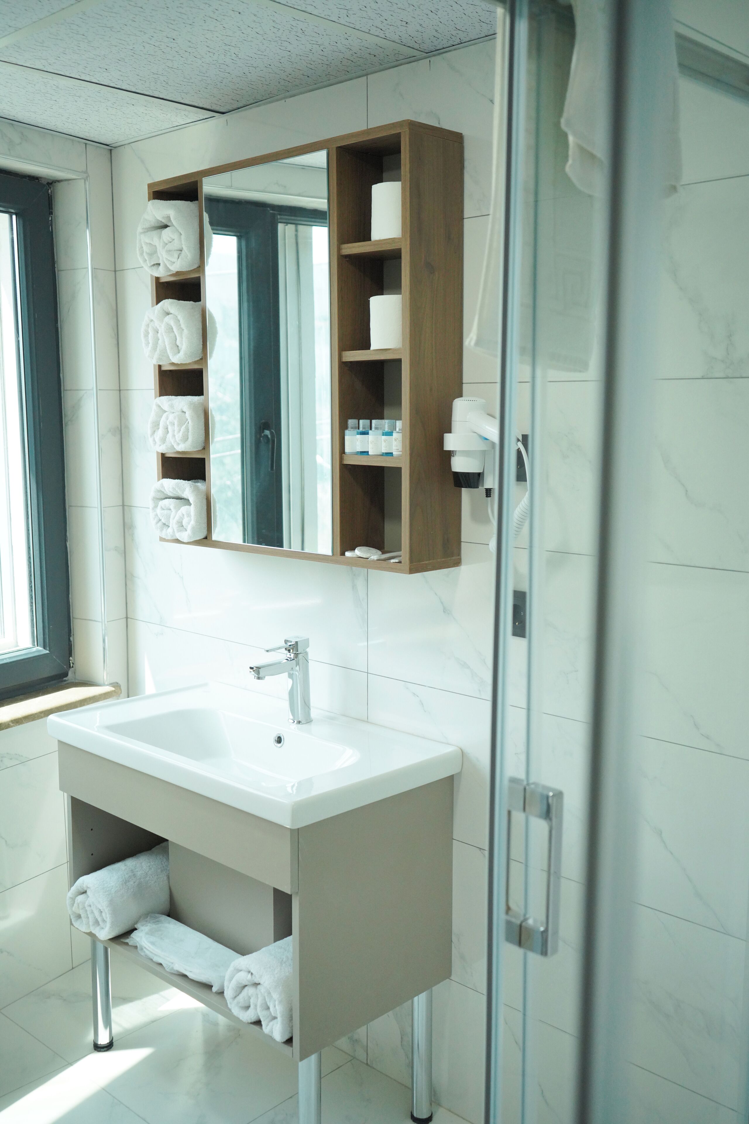 A modern bathroom with a marble floor, a white sink, a glass shower enclosure, and a wooden medicine cabinet above the sink.