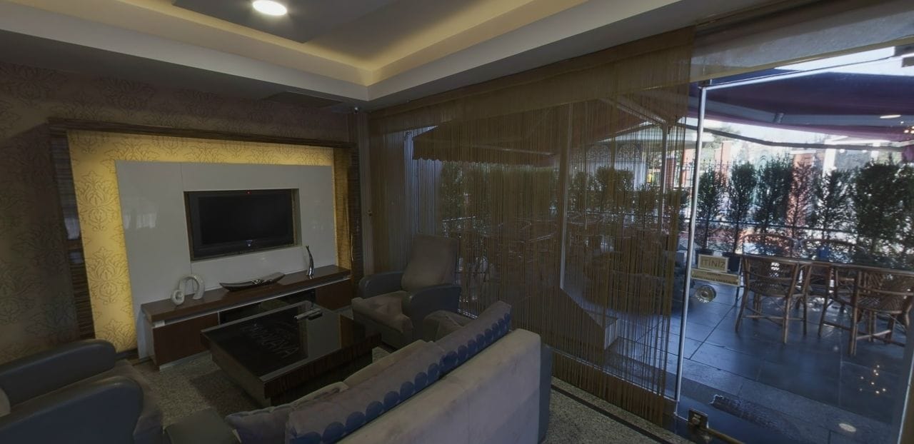 A living room with a flat-screen TV, a sofa, and a glass door leading to an outdoor patio with a view of trees and a building.