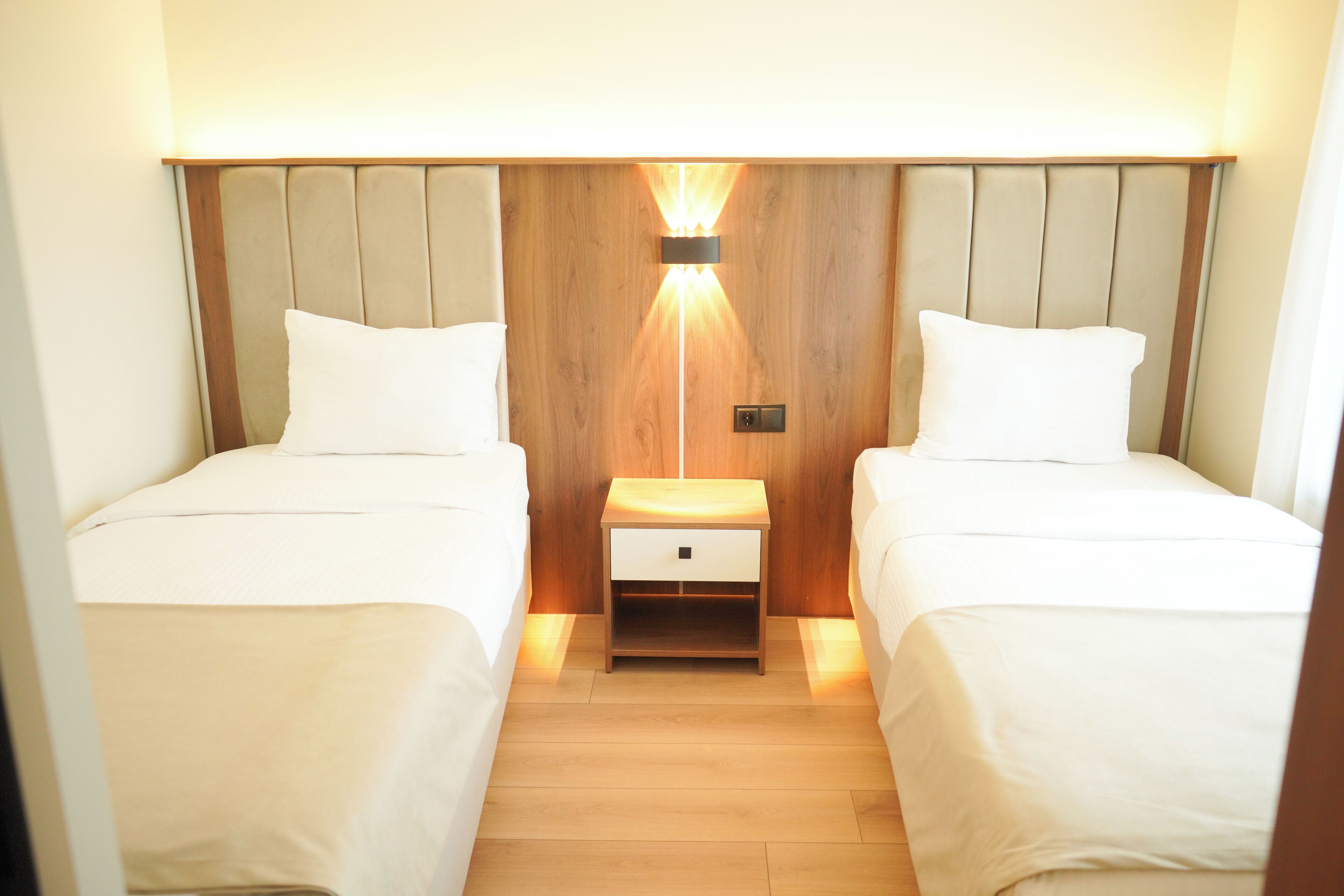 A hotel room with two beds, a wooden headboard, a nightstand with a lamp, and a small drawer.