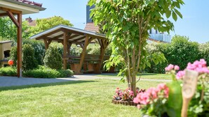A garden with a wooden pergola, a potted plant, and a building with a red roof in the background.