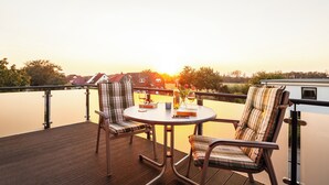 A deck with a round table and two chairs, overlooking a calm body of water and a row of houses.