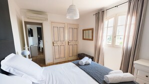 A hotel room with a bed, a bedside table, a lamp, a window with curtains, and a door leading to another room.