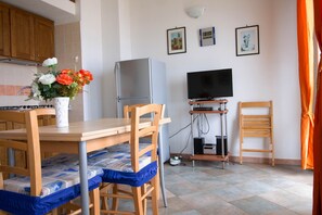 Eine kleine Küche mit Esstisch, Stühlen, einem Kühlschrank, einem Fernseher und einem an der Wand befestigten Regal.