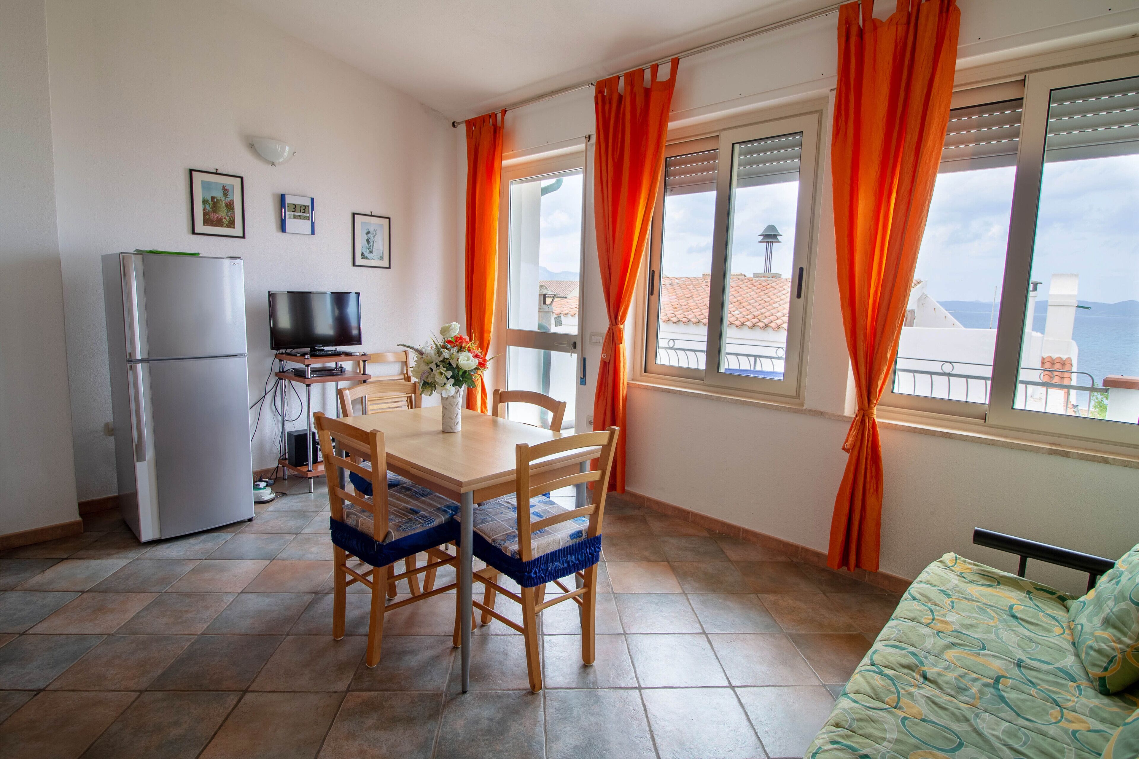 Eine kompakte Küche mit Kühlschrank, einem Esstisch mit Stühlen, einem Fernseher und einem Balkon mit orangenen Vorhängen.