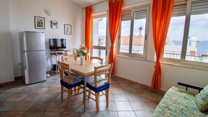 A compact kitchen with a refrigerator, a dining table with chairs, a TV, and a balcony with orange curtains. - House 100 meters from the sea Sardinia (SINISCOLA)