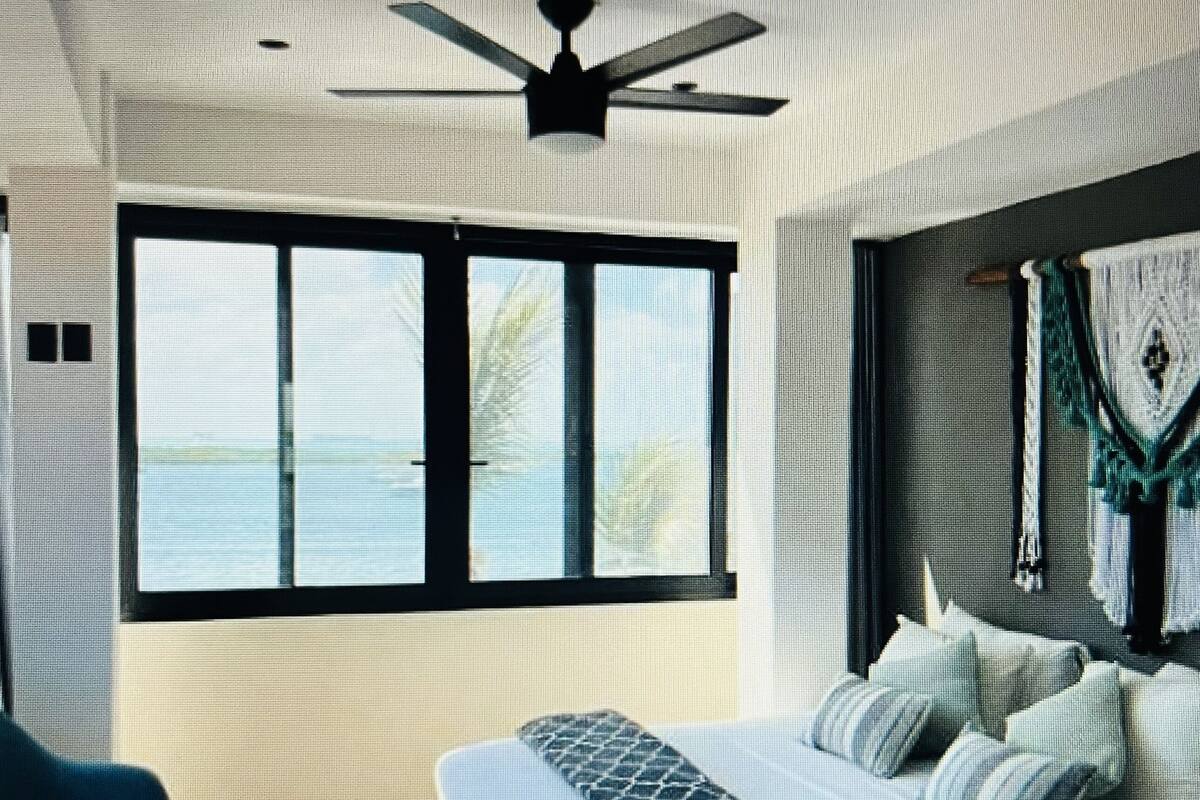 A modern bedroom with a large bed, a ceiling fan, and a view of the sea through a large window.