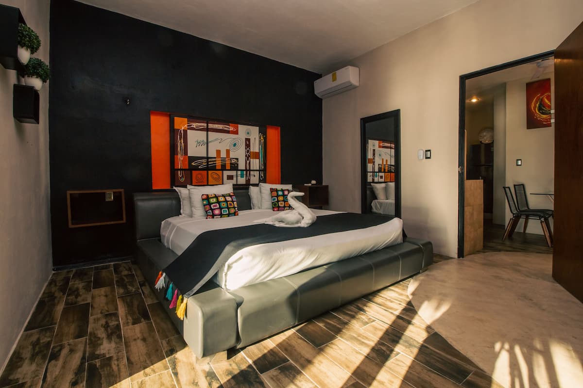 A modern bedroom with a large bed, a dark wall with a colorful abstract painting, and a small seating area with a chair.