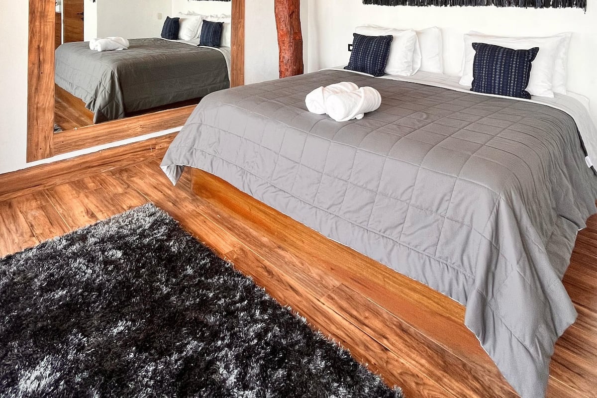A bedroom with a wooden ceiling, a large bed with grey bedding, a black and white wall hanging, and a mirror.