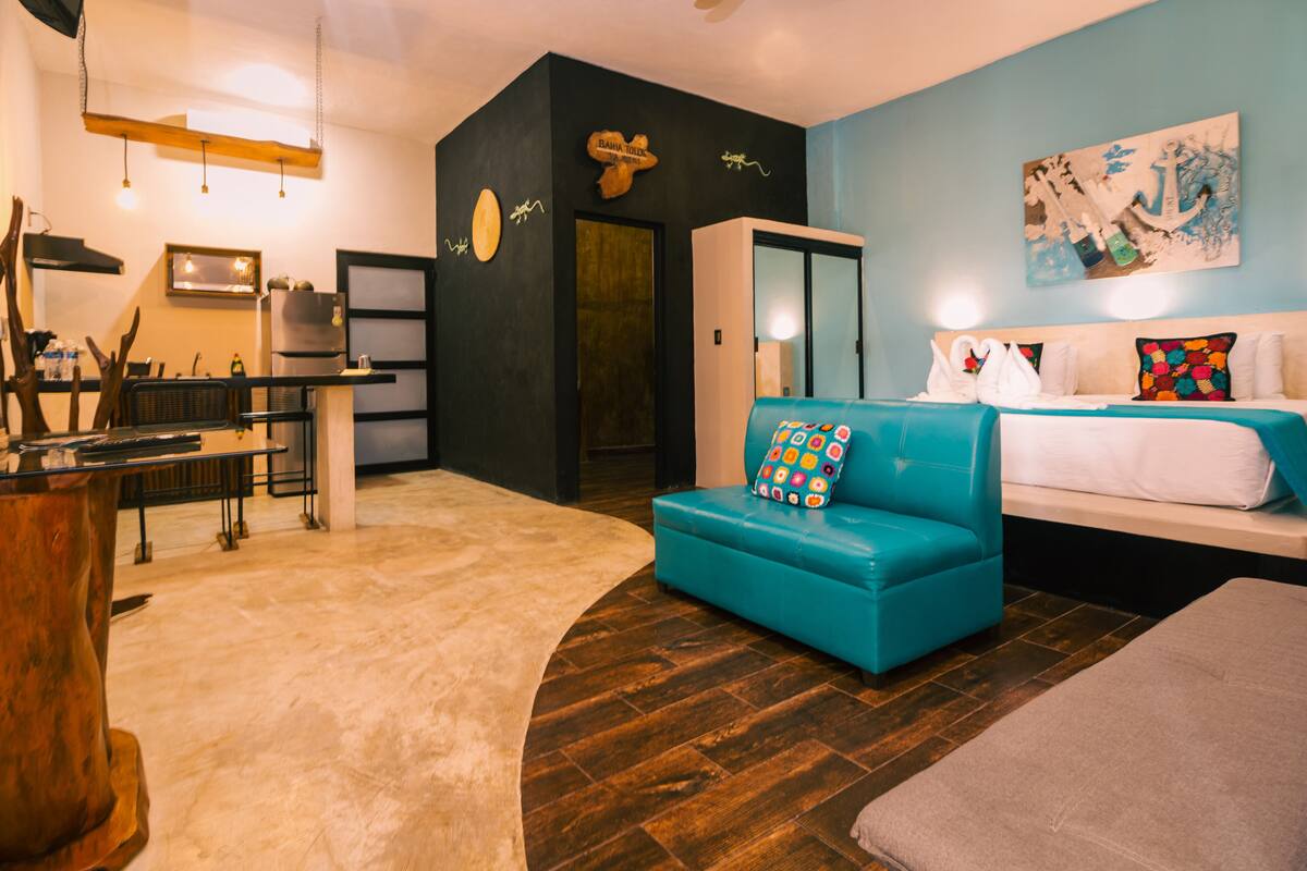A modern hotel room with a sofa, a bed, a dining area, and a kitchen.