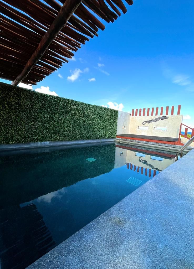 Una piscina cristalina con una pérgola de madera en lo alto, un seto verde y un muro rojo y blanco con una escultura.
