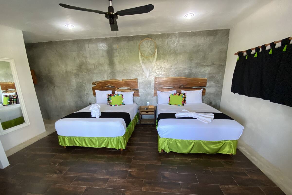 A hotel room with two beds, each with a wooden headboard, white bedding, and green skirts. There is a ceiling fan, a small table, and a mirror with a shelf of toiletries.