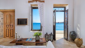 A room with a wooden table, a potted plant, a sofa, and two doors leading to a balcony with a view of the sea. - Halki Sea House (Halki)