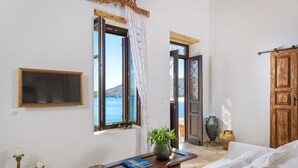 A room with a bed, a wooden table with a book and a vase, a wall-mounted TV, and a view of the sea through an open window. - Halki Sea House (Halki)