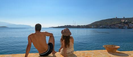 Dos personas sentadas en una terraza de piedra con vista a un cuerpo de agua y edificios al fondo.