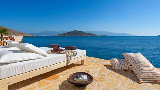 A sunlit outdoor seating area with a stone floor, a white sofa with pillows, a striped sofa, and a small table with a book and a decorative item. - Halki Sea House (Halki)