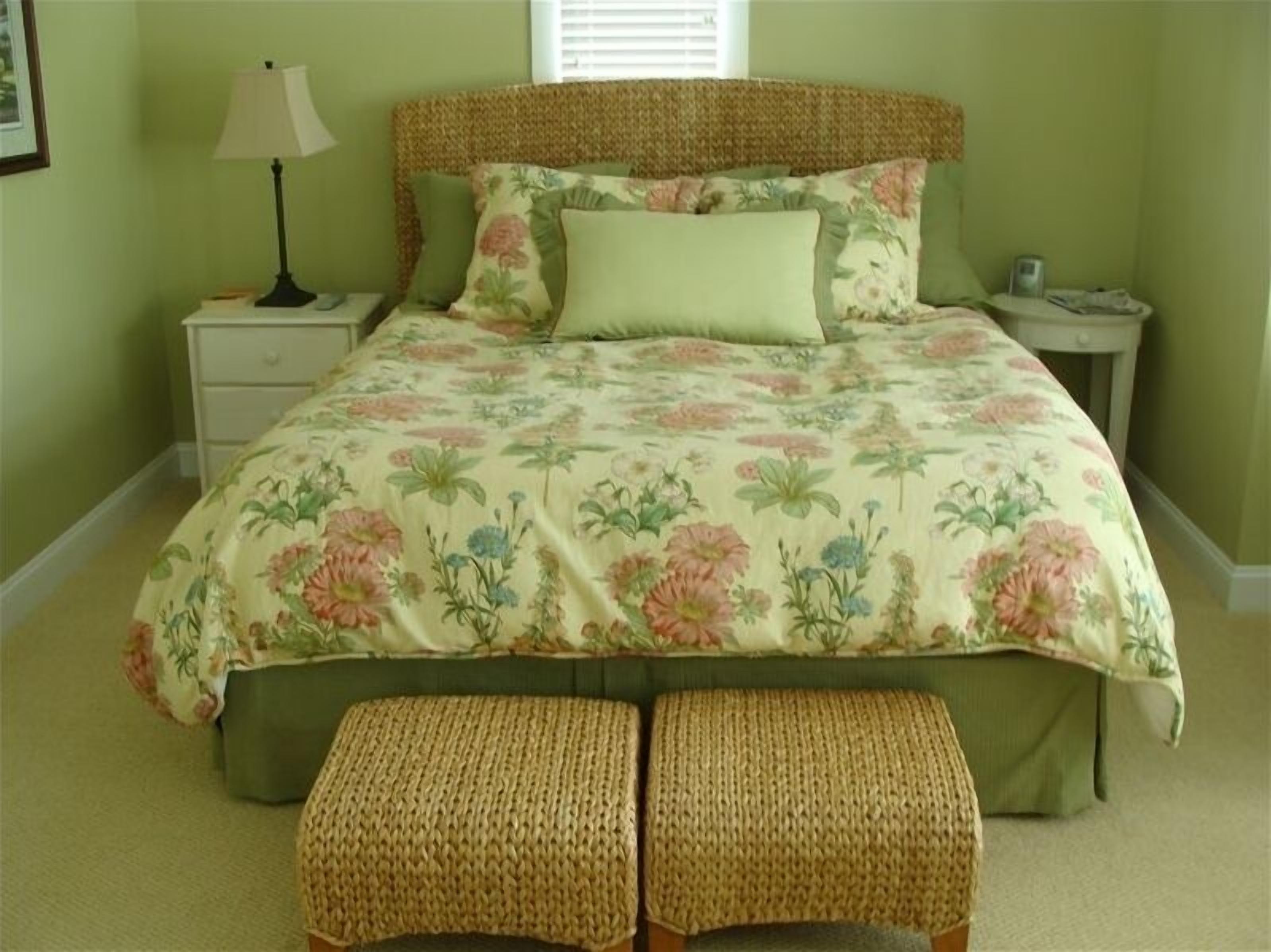 A bedroom with a floral bedspread, a woven bench, and a nightstand with a lamp.