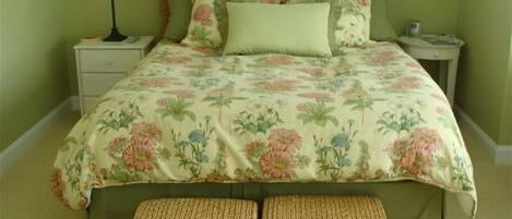 A bedroom with a floral bedspread, a woven bench, and a nightstand with a lamp.