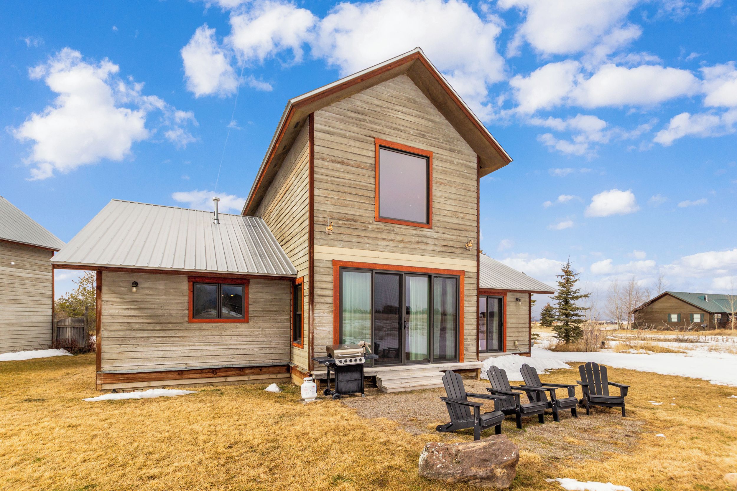 Une maison en bois à deux étages, dotée d’une grande porte-fenêtre coulissante, d’un barbecue et d’un espace extérieur aménagé avec des chaises.